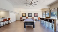 This single-floor amenity lounge is a well-appointed recreational space designed for social gatherings and leisure. The main living area commands attention with soaring vaulted ceilings clad in white shiplap and anchored by a large, dramatic ceiling fan. A full-size billiards table with a blue felt surface serves as the centerpiece of the room, surrounded by cognac leather wingback chairs and accented by bold, oversized billiard ball artwork lining the walls. Sputnik-style chandeliers add a mid-century modern flair throughout the space.

A striking, elongated bar island with a richly veined marble-look countertop and bar-height seating provides an ideal gathering spot, complementing the open and airy layout. Floor-to-ceiling windows and glass doors along one wall flood the space with natural light and offer views of the surrounding outdoor courtyard and building complex beyond.

The compact kitchenette is efficiently designed with white cabinetry, a sink, microwave, full-size refrigerator, and dishwasher — all in sleek black finishes — supporting event hosting needs. A hallway connects to a clean, simply appointed bathroom featuring neutral tones and curated wall art. Light wood-tone luxury vinyl plank flooring runs throughout, tying the entire space together with a cohesive, contemporary aesthetic.