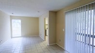 This single-floor space presents a straightforward and functional layout designed for comfortable everyday living. The main living area is finished with light-toned ceramic tile flooring that extends throughout, providing a clean and cohesive look underfoot. Warm beige walls create a neutral backdrop that complements a variety of furnishing styles, while white baseboards offer a crisp contrast throughout the space. The textured popcorn ceiling adds a classic touch overhead, and a smoke detector is centrally positioned in the living area. Natural light enters through a sliding glass door fitted with vertical blinds, offering both privacy and access to the outdoors. An open passageway connects the living area to the kitchen, which is visible in the background and features white cabinetry and standard appliances, including what appears to be a range. The open sightlines between rooms enhance the sense of flow and connectivity throughout the space. Overall, the layout is practical and move-in ready, with a simple, well-maintained aesthetic that provides a welcoming and livable environment.