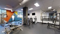This fitness facility is thoughtfully designed to accommodate a range of workout styles and fitness goals. The main exercise area features durable rubber flooring throughout, providing both safety and comfort during training sessions. A well-stocked dumbbell rack offers a variety of free weights, complemented by a weight bench for strength training exercises. Multiple elliptical machines are positioned centrally, offering users quality cardio options. Wall-mounted flat-screen televisions are strategically placed to keep members entertained during workouts. Natural light filters in through large windows on one side of the space, creating an energizing atmosphere, while recessed and panel ceiling lights ensure consistent illumination throughout. A bold, colorful geometric mural near the entrance adds a vibrant, motivating aesthetic to the space. A mirrored studio area with a ballet barre is situated along one wall, ideal for stretching, dance, or functional fitness classes. Structural columns are finished in a dark charcoal tone, providing a modern contrast to the clean white walls. The facility's open, single-floor layout maximizes usable space and allows for easy navigation between different training zones, making it a versatile and welcoming environment for fitness enthusiasts of all levels.