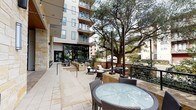 This expansive outdoor lounge is thoughtfully designed to offer a variety of seating experiences across a well-appointed patio space. The area is anchored by a striking natural limestone accent wall that lends a warm, organic character to the environment, complemented by rich wood-paneled ceiling details in the covered lounge section. Floor-to-ceiling bronze-framed glass walls visually connect the outdoor area to the interior, maximizing openness and light.

The covered lounge zone features plush all-weather sectional sofas and armchairs with crisp white cushions and sage green accent pillows, arranged around a wall-mounted flat-screen TV — ideal for relaxed gatherings. Adjacent to this is an open-air terrace centered around a round stone fire pit with a glass bead interior, encircled by additional wicker lounge seating with white cushions.

Further into the space, multiple dining sets with glass-top tables and wicker chairs are arranged beneath bistro-style string lights strung between sleek black poles. Horizontal wood privacy screens and black metal railings define the perimeter, while mature live oak trees provide natural shade and a lush backdrop. The light-toned stone tile paving runs throughout, unifying the entire lounge into a cohesive, resort-inspired outdoor retreat.