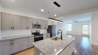 Located at 1588 Bloom St in Mount Pleasant, South Carolina, this single-floor home offers a clean, contemporary design with thoughtfully selected finishes throughout. The open-concept kitchen serves as the heart of the home, showcasing gray shaker-style cabinetry with matte black hardware, granite countertops, a classic white subway tile backsplash, and a full suite of stainless steel appliances. A generously sized kitchen island with an integrated sink and modern pendant lighting makes this space ideal for both cooking and entertaining. Light hardwood flooring flows seamlessly through the main living and kitchen areas, adding warmth and continuity. The living area benefits from natural light through a window and features a ceiling fan for added comfort. Bedrooms are carpeted and bright, each equipped with ceiling fans and ample closet access. The bathroom hallway features matching granite-topped white cabinetry with matte black hardware, leading to a walk-in glass-enclosed shower. A dedicated laundry room with full-size washer and dryer hookups and overhead cabinetry adds practical convenience. The overall aesthetic is modern and neutral, with crisp white walls and consistent design elements creating a cohesive, move-in-ready environment.
