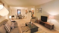 This single-floor residence offers an open-concept layout that seamlessly connects the living, dining, and kitchen areas, creating a cohesive and functional living environment. The living room serves as the central gathering space, featuring plush carpeting in warm neutral tones and a generous footprint accommodating a full sofa arrangement, accent chairs, and a sleek black coffee table atop a chevron-patterned area rug. A wood-toned media console with an mounted flat-screen television anchors one wall, while the layout allows for comfortable flow and conversation throughout the space. Adjacent to the living area, the dining space is defined by a pendant light fixture that adds a decorative focal point above the dining table, which is surrounded by upholstered chairs. Large windows in the dining area invite natural light and provide views to the exterior. A pass-through opening connects the dining area to the kitchen, which features warm wood cabinetry and standard appliances. The overall ambiance throughout the space is warm and inviting, with soft lighting, neutral wall tones, and consistent carpeting creating a unified and comfortable atmosphere.
