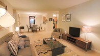 This single-floor residence offers an open-concept layout that seamlessly connects the living, dining, and kitchen areas, creating a cohesive and functional living environment. The living room serves as the central gathering space, featuring plush carpeting in warm neutral tones and a generous footprint accommodating a full sofa arrangement, accent chairs, and a sleek black coffee table atop a chevron-patterned area rug. A wood-toned media console with an mounted flat-screen television anchors one wall, while the layout allows for comfortable flow and conversation throughout the space. Adjacent to the living area, the dining space is defined by a pendant light fixture that adds a decorative focal point above the dining table, which is surrounded by upholstered chairs. Large windows in the dining area invite natural light and provide views to the exterior. A pass-through opening connects the dining area to the kitchen, which features warm wood cabinetry and standard appliances. The overall ambiance throughout the space is warm and inviting, with soft lighting, neutral wall tones, and consistent carpeting creating a unified and comfortable atmosphere.
