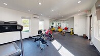 Located at 345 Bryant Rd in Spartanburg, SC, this single-floor fitness center offers a well-appointed workout space designed to support a variety of exercise routines. The room features a dark, rubberized flooring ideal for high-impact activities, crisp white walls, and a recessed lighting system that keeps the space bright and functional throughout the day. Multiple windows along the walls invite natural light and provide views of the surrounding greenery, contributing to an energizing atmosphere. A wall-mounted mini-split HVAC unit ensures year-round climate comfort during workouts. A large floor-to-ceiling mirror spans one wall, visually expanding the room and allowing users to monitor their form. The space is equipped with a rowing machine, spin bike, and treadmill for cardiovascular training, as well as a dumbbell rack with multiple weight options for strength work. Stability balls, medicine balls, and a yoga mat area cater to flexibility and functional fitness training. A flat-screen television is mounted on the wall for entertainment, and a wall-mounted sanitation station promotes cleanliness. The overall design is clean, modern, and practical, making this fitness center a capable and efficient facility for daily exercise.