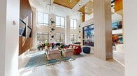 Located at 1733 Storrs Rd in Storrs Mansfield, CT, this expansive single-floor amenity space delivers a striking blend of contemporary design and vibrant social atmosphere. Soaring double-height ceilings anchor the main lounge area, where floor-to-ceiling windows flood the interior with abundant natural light and offer views of the surrounding landscape. Light wood-plank flooring runs throughout, complementing warm wood-paneled accent walls and crisp white surfaces.

The central lounge area features a bold fireplace framed in richly veined stone, with a flat-screen TV mounted above and plush seating arrangements anchored by teal and orange furnishings. Adjacent to this, a games area hosts a full-size billiards table beneath pendant lighting, alongside a ping-pong table and a navy-accented bar with counter seating.

Intimate arched alcoves provide semi-private booth seating with teal upholstery, slatted wood paneling, and patterned wallcoverings, ideal for smaller gatherings. A welcoming reception area near the entrance features leather lounge chairs, statement pendant lighting, and a staffed desk beneath a wall of tall windows.

Curated artwork, including large-scale murals and sculptural wall installations, is displayed throughout. The elevator lobby showcases polished marble-effect wall cladding, completing this thoughtfully designed, amenity-rich community space.