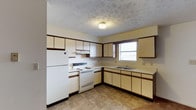 Katelyn Court Apartments presents a practical and well-organized single-floor residence designed for comfortable everyday living. The kitchen is a standout feature of the unit, showcasing an L-shaped layout that maximizes efficiency and usability. Cream-colored cabinetry with dark wood trim accents lines both the upper and lower walls, providing generous storage throughout. The kitchen is equipped with a full-size white refrigerator, an electric range with cooktop, and a built-in double sink positioned beneath a window that allows natural light to brighten the space. A ventilation hood is installed above the range, and additional upper cabinets offer plenty of room for pantry storage. The flooring features a durable tile pattern in warm earthy tones, adding a classic, easy-to-maintain surface underfoot. A textured ceiling with a decorative embossed finish adds a subtle design detail overhead. The overall ambiance of the space is clean, functional, and straightforward, making it a solid option for those seeking a no-fuss residential setting with all the essential amenities in place.