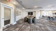 Located at 30000 Summer Crop Lane in Apex, NC, this generously sized single-floor community space offers a thoughtfully designed layout that seamlessly integrates a dining room, living room, and lounge areas within one cohesive floor plan. The interior design is contemporary and refined, featuring a neutral palette of warm beiges, soft grays, and crisp whites throughout, complemented by recessed lighting and tray ceiling details that add architectural depth to each area.

The dining area accommodates multiple seating arrangements, with round marble-topped tables paired with modern upholstered chairs arranged across a richly patterned carpet. A built-in bar station with dark cabinetry and counter seating anchors one end of the room, providing a functional and stylish gathering point. Curated wall art and decorative accents are thoughtfully placed throughout.

The living room area features a fireplace flanked by built-in shelving, a wall-mounted television, and plush seating arrangements that create an inviting atmosphere. A dedicated lounge nook with a round gaming table and barrel-style chairs offers an additional social setting.

Outdoor balconies with black metal railings provide open-air access, offering elevated views of the surrounding courtyard and landscaped community grounds beneath a wide, open sky.