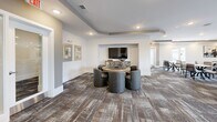 Located at 30000 Summer Crop Lane in Apex, NC, this generously sized single-floor community space offers a thoughtfully designed layout that seamlessly integrates a dining room, living room, and lounge areas within one cohesive floor plan. The interior design is contemporary and refined, featuring a neutral palette of warm beiges, soft grays, and crisp whites throughout, complemented by recessed lighting and tray ceiling details that add architectural depth to each area.

The dining area accommodates multiple seating arrangements, with round marble-topped tables paired with modern upholstered chairs arranged across a richly patterned carpet. A built-in bar station with dark cabinetry and counter seating anchors one end of the room, providing a functional and stylish gathering point. Curated wall art and decorative accents are thoughtfully placed throughout.

The living room area features a fireplace flanked by built-in shelving, a wall-mounted television, and plush seating arrangements that create an inviting atmosphere. A dedicated lounge nook with a round gaming table and barrel-style chairs offers an additional social setting.

Outdoor balconies with black metal railings provide open-air access, offering elevated views of the surrounding courtyard and landscaped community grounds beneath a wide, open sky.
