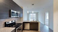 Fifth Street Place offers a thoughtfully designed single-floor residence that seamlessly blends modern aesthetics with functional living. The open-concept layout connects the kitchen and living area, creating a cohesive and airy environment enhanced by neutral tones and light wood-style flooring throughout.

The kitchen is a standout feature, showcasing dark wood-grain cabinetry that extends to the upper level for generous storage. A peninsula with granite countertops and an integrated sink acts as a natural divider between the kitchen and living space, while also providing casual seating or prep space. Stainless steel appliances, including a range, microwave, and dishwasher, complement the sleek gray subway tile backsplash.

Two pendant lights hang above the peninsula, adding focused illumination and a contemporary design touch. The living area beyond benefits from a large window that draws in natural light and offers views of the surrounding greenery, contributing to a bright and welcoming ambiance. The clean white walls and open sightlines reinforce the sense of spaciousness, making the overall space feel modern, practical, and inviting.