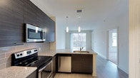 Fifth Street Place offers a thoughtfully designed single-floor residence that seamlessly blends modern aesthetics with functional living. The open-concept layout connects the kitchen and living area, creating a cohesive and airy environment enhanced by neutral tones and light wood-style flooring throughout.

The kitchen is a standout feature, showcasing dark wood-grain cabinetry that extends to the upper level for generous storage. A peninsula with granite countertops and an integrated sink acts as a natural divider between the kitchen and living space, while also providing casual seating or prep space. Stainless steel appliances, including a range, microwave, and dishwasher, complement the sleek gray subway tile backsplash.

Two pendant lights hang above the peninsula, adding focused illumination and a contemporary design touch. The living area beyond benefits from a large window that draws in natural light and offers views of the surrounding greenery, contributing to a bright and welcoming ambiance. The clean white walls and open sightlines reinforce the sense of spaciousness, making the overall space feel modern, practical, and inviting.