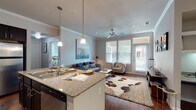 This single-floor residence features a thoughtfully designed open-concept layout that seamlessly connects the kitchen, dining, and living areas. The kitchen is outfitted with dark espresso cabinetry, stainless steel appliances including a range and microwave, and a granite-topped island with bar seating illuminated by pendant lighting. Track lighting and crown molding add refined detail throughout.

The living area extends naturally from the kitchen, featuring rich hardwood-style flooring, crown molding, a ceiling fan, and multiple large windows fitted with blinds that allow generous natural light. A dedicated seating nook near the windows offers additional versatility.

Two bedrooms provide comfortable accommodations, each with ceiling fans, carpeted floors, ample natural light from multiple windows, and walk-in or standard closets. The primary bedroom includes a private en-suite bathroom featuring a granite vanity, dark cabinetry, tiled tub-shower combination, and tile flooring. A second full bathroom mirrors this design with similarly clean, neutral finishes.

A dedicated laundry closet houses a full-size washer and dryer. Throughout, the unit maintains a consistent palette of neutral tones, granite surfaces, and dark-finished cabinetry, contributing to a cohesive and functional living environment.
