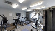 This single-floor fitness center is designed to support a well-rounded workout experience. The space is outfitted with a range of cardio equipment, including a recumbent bike, an elliptical trainer, and a treadmill, providing users with multiple options for aerobic exercise. A multi-station cable machine anchors the strength training area, complemented by a full rack of dumbbells organized neatly along the wall for easy access. The flooring features a combination of patterned carpet and rubberized exercise mats, balancing comfort with functionality across different workout zones. The walls are finished in a clean, neutral gray, contributing to a focused and energizing atmosphere. Overhead, flush-mounted fluorescent light panels provide bright, even illumination throughout the room, while a large arched window allows natural light to fill the space, enhancing the overall ambiance. A wall-mounted television is positioned to keep exercisers entertained during cardio sessions. The layout is thoughtfully arranged to allow adequate space between equipment, ensuring safe and comfortable movement throughout the facility.