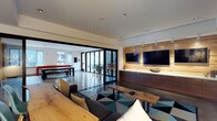 This single-floor space is designed with a contemporary, open-plan layout that connects multiple functional areas in a cohesive and visually engaging environment. The main lounge area is anchored by a plush sectional sofa paired with a live-edge wood coffee table and a bold geometric-patterned area rug in teal, navy, and charcoal tones, adding a striking design focal point to the space.

Along one wall, a built-in media unit crafted from rich dark wood cabinetry features a white countertop surface and warm under-cabinet lighting. Two flat-screen televisions are mounted side-by-side above, complemented by a rustic wood accent panel with track lighting overhead, creating a warm contrast against the clean white walls and ceiling.

Recessed lighting throughout maintains a bright and modern ambiance. Large black-framed sliding glass doors open directly to an adjacent area furnished with bar-height seating and a long dining table, allowing natural light to flow freely between spaces and offering a seamless transition for gatherings. The polished concrete-style flooring unifies the entire floor plan, reinforcing the space's sleek, modern aesthetic throughout.