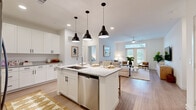 This thoughtfully designed 2-bedroom, 2-bathroom residence in Austin, TX features a clean, modern aesthetic throughout its single-story open-concept layout. The kitchen serves as a focal point of the home, showcasing white shaker cabinetry, granite countertops, stainless steel appliances—including a French door refrigerator, range, and built-in microwave—and a large center island with an undermount sink and seating for multiple guests, illuminated by three bold black pendant lights. Light wood-tone flooring runs throughout the main living areas, lending warmth and continuity. The adjoining living room is generously proportioned, featuring a ceiling fan, large windows with French doors that invite abundant natural light and outdoor views, creating a bright and welcoming atmosphere. Both bedrooms are comfortably sized, each benefiting from natural light, and are accompanied by dedicated closets. The primary bedroom connects to its own bathroom, which features granite-topped white cabinetry, a framed mirror, and open shelving. The second bathroom mirrors the same refined finishes. Together, the spaces deliver a cohesive, contemporary feel well-suited to modern living in the heart of Austin.