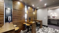 This single-floor space showcases a striking blend of rustic and contemporary design elements. The most prominent feature is the rich, horizontal wood plank paneling that lines the walls, lending warmth and texture to the interior. The natural tones of the wood create a welcoming atmosphere while providing a bold visual backdrop throughout the space.

Tall, narrow vertical light fixtures are integrated between the wood panels, offering ambient illumination that complements the recessed ceiling lights above. The combination of these lighting sources ensures a well-lit environment while maintaining a cozy, intimate feel.

The seating area is furnished with sturdy wooden bar-height tables paired with industrial-style stools featuring round wooden seats and black metal bases. This furniture arrangement encourages casual interaction and is well-suited for communal use or collaborative gatherings.

The flooring features a patterned carpet in muted grey and charcoal tones, contrasting effectively with the warm wood tones above. A decorative metal sculpture serves as a tabletop accent, adding an artistic touch to the space. Overall, the design balances rustic warmth with modern industrial elements, resulting in a functional yet aesthetically refined environment.