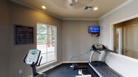 Located at 155 Lakeside Dr in Waxahachie, TX, this single-floor fitness center offers a well-equipped workout space designed for both cardio and strength training. The room features neutral gray walls accented by classic crown molding and is finished with dark rubber flooring, providing both durability and comfort underfoot. Recessed lighting and a ceiling fan ensure the space remains well-lit and well-ventilated during workouts.

The facility is equipped with a treadmill, elliptical trainer, stationary bike, and a multi-station cable strength machine, catering to a full range of fitness routines. A wall-mounted flat-screen television adds entertainment for those engaging in longer cardio sessions. A full-length mirror along one wall visually expands the space and allows users to monitor their form during exercise.

Natural light flows in through large windows, including two prominent windows that frame a direct view of the outdoor pool area, complete with palm trees, lounge seating, and a pergola — creating an inviting and motivating atmosphere. The space is accessed through a charming wood-framed French door with glass pane inserts. Fitness center rules are posted prominently, reflecting a well-managed, community-oriented facility.