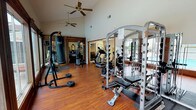 This fitness center is designed to support a comprehensive workout experience within a thoughtfully arranged, single-level space. The main workout area features soaring vaulted ceilings accented by dual ceiling fans, providing ample airflow and an open, airy atmosphere. Rich hardwood-style flooring runs throughout, lending warmth and durability underfoot.

The strength training zone is anchored by a full cable machine and smith rack system, complemented by free weights, a weight bench, and a freestanding heavy bag for boxing and functional training. Large floor-to-ceiling windows along one wall frame direct views of an outdoor pool area, filling the space with natural light and creating a motivating backdrop during workouts.

The cardio section includes a True treadmill, an elliptical trainer, and an exercise bike, positioned to take advantage of both the natural light and the pool views. Wall-mounted mirrors throughout the facility enhance the sense of space and allow users to monitor their form. An adjoining room provides additional cardio equipment, protected by cushioned floor mats, and features a full-wall mirror and a bulletin board for community notices. The overall ambiance is clean, functional, and inviting, suited for a variety of fitness routines.