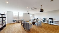 This single-floor multipurpose room offers a well-organized layout that combines several functional zones within a generous open footprint. The space is finished with warm golden-toned carpeting and light gray walls, creating a welcoming and neutral aesthetic throughout.

Natural light is a defining feature, with large windows along one wall providing views of surrounding treetops and flooding the interior with daylight. The ceiling is fitted with recessed lighting and ceiling-mounted HVAC units, complemented by stylish bronze pendant lights that add a refined decorative element to the space.

The lounge area features upholstered armchairs with geometric patterned fabric arranged around small circular side tables, offering a comfortable setting for casual gathering or reading. Adjacent to the lounge, a dedicated computer workstation zone includes a long white desk lined with desktop computers and ergonomic office chairs, supporting productivity and digital access.

A full bookshelf unit along one wall serves as an in-room library, stocked with a wide variety of books and adding both visual interest and functional value. Multiple white-paneled doors along the back wall provide access to additional storage or adjoining spaces. The overall ambiance is modern yet comfortable, balancing communal utility with a relaxed, residential feel.