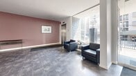 Located at 1865 N Fuller Ave in the heart of Los Angeles, this building lobby presents a clean, modern aesthetic designed to make a strong first impression. The space is finished with large-format stone tile flooring in a cool grey tone, lending a polished and professional feel underfoot. A seating area near the expansive floor-to-ceiling glass façade features sleek black leather armchairs arranged around a small accent table, bathed in abundant natural light with views of the surrounding courtyard and building exterior. The glass entry doors open to a welcoming foyer complete with a grey entrance mat, while a decorative stone-clad column adds a tactile architectural accent. Crisp white walls are complemented by a warm mauve accent wall, and framed artwork adds a curated touch. A stainless steel elevator provides vertical access within the building. Recessed ceiling lighting ensures even illumination throughout. Toward the rear of the lobby, a dedicated mailroom alcove with individual mailboxes and wall sconce lighting offers a functional, well-organized service area. A live potted tree near the entrance adds a refreshing natural element. Overall, the lobby strikes a balance between contemporary design and practical functionality.