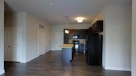 This single-floor residence offers a thoughtfully designed open-concept layout that seamlessly blends the kitchen and living areas into one cohesive, functional space. The kitchen is a standout feature, showcasing rich dark cabinetry that extends to the upper walls, providing ample storage while creating a bold, contemporary aesthetic. A central island with a light granite countertop serves as both a prep surface and casual gathering spot, complemented by stylish pendant lighting that adds warmth and visual interest overhead. Black appliances, including a refrigerator, range, and built-in microwave, complete the kitchen's sleek, modern look. The flooring throughout is a rich, dark hardwood-style material that anchors the space with warmth and continuity. Neutral light gray walls keep the interior feeling open and airy, allowing the darker design elements to take center stage without overwhelming the space. Flush-mount ceiling lighting provides consistent illumination across the living area, while the smooth, clean-lined ceilings contribute to the overall sense of openness. Closet and utility access doors are neatly integrated into the design, maintaining a tidy and uncluttered appearance. The overall ambiance is modern and inviting, with a well-balanced mix of dark and light tones.