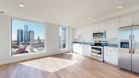 This single-floor unit showcases a contemporary design defined by clean lines, crisp white walls, and light wood flooring that runs consistently throughout the space. Large floor-to-ceiling windows are a standout feature, flooding every room with abundant natural light while framing striking panoramic views of the surrounding urban skyline, including notable downtown landmarks and cityscape vistas.

The open-concept kitchen is well-appointed with white shaker-style cabinetry extending to the ceiling, stone-look countertops, and a full suite of stainless steel appliances including a refrigerator with ice and water dispenser, electric range, and built-in microwave. The kitchen connects seamlessly to the living area, enhancing the sense of openness.

Multiple bedrooms offer generous proportions, each benefiting from large windows with city views and closets with built-in shelving for organized storage. The bathroom features a wood-toned vanity with a stone-finish countertop and clean, modern fixtures. The entry hallway is functional and well-lit, featuring sleek modern door hardware. Recessed lighting throughout complements the abundant natural light, contributing to an overall bright and airy atmosphere.