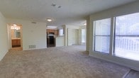 This single-floor residence offers a straightforward and functional layout designed for comfortable everyday living. The main living area is spacious and open, featuring wall-to-wall carpeting in a neutral tone that adds warmth underfoot. Large windows dressed with horizontal blinds allow abundant natural light to fill the space, creating a bright and airy atmosphere throughout.

The kitchen is situated adjacent to the living area and is equipped with black appliances, including a range and microwave, set against warm wood-toned cabinetry. A light-colored peninsula counter serves as a natural divider between the kitchen and the main living space, offering additional prep or seating options.

Beyond the living area, a secondary space — likely a bedroom or flex room — is visible and features built-in shelving or closet storage with louvered doors, also benefiting from its own window for natural light. The textured ceiling and neutral beige walls are consistent throughout, giving the home a cohesive and clean aesthetic. Overhead lighting fixtures provide practical illumination in key areas. The overall layout is open and efficient, making good use of the available floor space across a single level.