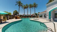 Located at 15645 N 35th Ave in Phoenix, Arizona, this single-level outdoor amenity space delivers a resort-style experience designed for relaxation and community gathering. At the heart of the space is a generously sized, freeform swimming pool with crystal-clear turquoise water, complemented by an attached spa. The pool deck is finished in light-toned concrete and is lined with teal market umbrellas shading multiple seating and dining arrangements, ideal for enjoying Arizona's sunny climate. Towering palm trees frame the area, adding a lush, tropical character against the backdrop of vivid blue desert skies. Adjacent to the pool, a clubhouse building features a distinctive teal arched entryway with large windows and a terra cotta tile roofline, providing a stylish architectural focal point. A covered outdoor lounge area, branded as "The Grove," offers a social retreat complete with turf flooring, teal and blue cushioned seating, string lighting, and a fire pit centerpiece — perfect for evening gatherings. A dedicated BBQ station nearby supports outdoor cooking and entertaining. A separate gated patio along the building's exterior provides additional seating with a shade umbrella, offering a quieter outdoor retreat. Together, these amenities create a vibrant and well-appointed outdoor environment.