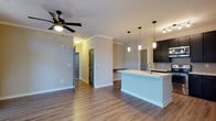 Located at 11 Parklands Drive in Bluffton, South Carolina, this single-story residence offers a thoughtfully designed open-concept floor plan that seamlessly connects the main living areas. The space is finished with warm hardwood-style flooring that flows consistently throughout, contributing to a cohesive and inviting atmosphere.

The kitchen is a standout feature, showcasing dark espresso cabinetry paired with a light-toned countertop island that doubles as a breakfast bar. A classic subway tile backsplash adds texture and contrast, while stylish pendant lights suspended above the island provide focused task lighting and a modern decorative touch. The open layout ensures the kitchen remains fully integrated with the surrounding living space, making it ideal for both everyday living and entertaining.

Crown molding along the ceilings adds a refined architectural detail throughout the home. A ceiling fan with a matte black finish provides both function and style in the main living area. Interior doors are finished in a neutral gray tone, complementing the light walls and overall contemporary color palette.

A hallway connects the living areas to additional rooms, with louvered closet doors visible along the corridor, offering practical storage solutions. The space presents a clean, move-in-ready aesthetic with modern fixtures and a neutral palette that allows for personalization.