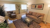 This single-floor space centers around a bright and inviting living room that combines functionality with a warm, homey ambiance. The room features polished parquet-style tile flooring that reflects natural light streaming in through multiple windows fitted with white blinds, creating an airy and open atmosphere throughout.

The living area is generously sized, comfortably accommodating a full sectional sofa with decorative accent pillows alongside a matching chaise lounge chair, both in a neutral taupe tone. A glass-top coffee table with a dark metal frame sits atop a rich, traditionally patterned area rug, adding a layer of texture and warmth to the space. A tall floor lamp and ceiling-mounted light fixture ensure the room is well-lit at any time of day.

Framed artwork on the walls adds a tasteful decorative touch, while a small bistro-style table and chairs positioned near the windows provide a casual secondary seating or dining nook. The clean, neutral wall tones and polished flooring contribute to a cohesive and well-maintained aesthetic, making the space feel both practical and welcoming.