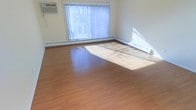 This single-floor space offers a clean, functional design with an open and airy feel. The main living area is finished with warm-toned laminate hardwood flooring that reflects natural light, creating a welcoming atmosphere throughout. A large picture window dressed with vertical blinds serves as the focal point of the room, allowing generous amounts of natural light to flood the interior and casting a bright, cheerful ambiance across the space. Baseboard heating units run along the perimeter of the room, ensuring efficient and consistent warmth throughout the colder months. The walls are finished in a neutral white tone, providing a versatile backdrop that complements a variety of interior styles and furnishing choices. The open layout of the main area offers flexibility for multiple uses, whether as a living room, dining space, or home office. A wall-mounted unit is also present, adding to the room's practical features. Overall, the space is characterized by its simplicity, brightness, and functional design, making it well-suited for a wide range of occupants looking for a comfortable and adaptable environment.