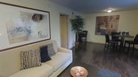 This single-floor residence offers a thoughtfully designed open-concept layout that seamlessly connects the living and dining areas, creating a cohesive and inviting atmosphere. Rich, dark hardwood flooring extends throughout the main living space, adding warmth and a sense of sophistication to the interior.

The living area is anchored by a comfortable cream sofa accented with decorative throw pillows in navy and neutral tones, complemented by a low-profile coffee table. Large framed abstract artwork adorns the walls, contributing to the space's contemporary aesthetic. A decorative bowl with sculptural accents serves as a refined focal point on the coffee table.

The dining area, visible from the living space, features a dark bistro-style table and chairs, creating an intimate setting for meals. A tall decorative indoor plant adds a natural, organic element to the space, bridging the living and dining zones with a touch of greenery.

The interior is finished with light-toned walls that contrast beautifully against the deep ceiling and flooring, enhancing the overall depth and dimension of the space. A doorway leads to an adjacent room, suggesting additional private living quarters. The combination of modern artwork, curated décor, and quality finishes gives this residence a polished, move-in-ready character.