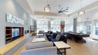 This single-floor clubhouse is designed as a welcoming communal gathering space with a thoughtfully open layout. The main lounge area is expansive, featuring elevated tray ceilings with recessed lighting and large industrial-style ceiling fans that enhance the sense of height and airflow. Pendant drum light fixtures hang at varying levels, adding a polished, modern aesthetic throughout.

The seating area is generously furnished with plush navy-blue sofas and complementary gray accent couches, arranged around low-profile stone-topped coffee tables to encourage both relaxed lounging and social interaction. A built-in fireplace anchors one wall, flanked by a decorative open shelving unit displaying curated accessories. Adjacent to the lounge, a billiards table provides a recreational focal point, further enhancing the clubhouse's entertainment offerings.

Light hardwood flooring runs throughout, contributing warmth and continuity to the space. A wall of large windows dressed with floor-to-ceiling gray curtains floods the interior with natural light and offers views to the exterior. The overall ambiance is contemporary, airy, and inviting — well-suited for community gatherings and leisure activities.