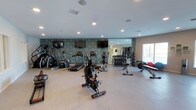 This fitness center offers a comprehensive workout environment designed to accommodate a wide range of exercise routines. The space is anchored by a row of cardio machines, including treadmills, ellipticals, a stair climber, and a stationary spin bike, arranged to allow comfortable use and movement throughout the room. A dedicated free weights area features a tiered dumbbell rack stocked with a full range of weights, accompanied by a workout bench and additional strength training accessories. A large wall mirror expands the sense of space and allows users to monitor their form during exercise. The accent wall showcases a bold, geometric patterned wallcovering in teal and neutral tones, adding a distinctive visual element to the room's atmosphere. Multiple flat-screen televisions are mounted along the feature wall, keeping users entertained during their workouts. Recessed lighting illuminates the space evenly from above, while a window along one wall allows natural light to filter in. The light-toned flooring and clean white ceiling contribute to a bright, open ambiance. Overall, the fitness center is thoughtfully laid out to balance both cardio and strength training needs in a well-appointed, inviting setting.