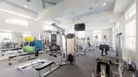 This fitness center offers a comprehensive and well-appointed workout environment designed to accommodate a wide range of exercise needs. The space is laid out across a single floor, with distinct zones dedicated to cardio training, strength machines, and free weights, allowing for an organized and efficient workout experience.

The architectural design is clean and modern, featuring tray ceilings with recessed lighting and multiple ceiling fans that ensure comfortable air circulation throughout. Large windows and glass-paneled doors flood the interior with abundant natural light, creating a bright and energizing atmosphere. The neutral color palette of whites and soft grays contributes to an open, airy feel.

The cardio area is equipped with multiple treadmills, ellipticals, and stationary machines, each featuring individual display screens for entertainment. The strength training section includes a robust selection of cable and resistance machines, as well as a dedicated free weights area stocked with a full rack of dumbbells, benches, and stability balls. Wall-to-wall mirrors in the free weights zone enhance the sense of space while providing functional visual feedback during workouts. Large flat-screen televisions are mounted prominently for viewing throughout the facility. Dark wood-style flooring ties the entire space together, delivering a polished and professional finish.