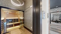 Located in El Paso, TX, this well-appointed fitness facility offers a comprehensive wellness experience across a single, generously sized floor. The main exercise room impresses with soaring ceilings, large floor-to-ceiling windows that flood the space with natural light and outdoor views, and a bold color-blocked carpet that adds visual depth. This area is outfitted with a full suite of Hoist strength machines, free weights on tiered racks, cable systems, benches, and cardio equipment including treadmills and ellipticals. A patterned accent wall adds a contemporary design touch, while ceiling fans, recessed lighting, and wall-mounted televisions enhance comfort and functionality. A built-in shelving unit with storage cabinetry provides additional organizational utility. The second exercise room serves as a dedicated cycling and functional training studio, featuring sage green walls, warm tray ceiling lighting, light hardwood flooring, full-length mirrors, spin bikes, and medicine ball racks — creating an intimate, focused environment. A glass-enclosed infrared sauna, finished in warm light wood paneling with tiered bench seating and a digital control panel, provides a premium recovery option. The facility is completed by a bathroom, rounding out the space as a fully self-contained wellness destination.