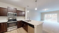 Located in Lakeville, MN, this well-appointed one-bedroom, one-bathroom residence offers a thoughtfully designed layout across a single floor. The open-concept kitchen and living area serves as the heart of the home, featuring rich dark-brown cabinetry, light stone-look countertops, a peninsula with a double-basin sink, pendant lighting, and a full suite of stainless steel appliances including a refrigerator, range, microwave, and dishwasher. Light hardwood-style flooring runs through the kitchen, transitioning to plush carpet in the living area and bedroom. The living room is bathed in natural light and offers access to a private balcony through sliding glass doors, overlooking a lush, natural landscape with mature trees. The spacious primary bedroom features generous closet space with bifold doors, a large window framing outdoor greenery, and soft carpeting throughout. The bathroom includes a wide vanity with dark cabinetry, a white countertop, a full-length mirror, and ample built-in storage. A connecting hallway provides additional closet space and seamless access between all rooms. Neutral wall tones and clean finishes throughout give the space a bright, move-in-ready feel.