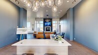 This contemporary single-floor clubhouse is designed to serve as a vibrant community gathering space, combining stylish aesthetics with practical amenities. The entrance foyer greets visitors with floor-to-ceiling glass doors that flood the space with natural light and offer views of the surrounding landscape.

At the heart of the space is a sleek reception desk adorned with a striking cluster of globe pendant lights in brass and glass, setting an upscale tone. The walls are finished in a calming steel-blue palette that carries throughout much of the space.

The expansive open lounge area is a standout feature, anchored by a dramatic floor-to-ceiling fireplace clad in dark veined marble, topped by a large wall-mounted television. Generous windows on multiple sides bring in abundant natural light. Comfortable tan leather seating and a low-profile wood coffee table create a welcoming social environment. Geometric LED chandelier fixtures overhead add a modern architectural touch.

Transitioning to the back of the space, a dedicated game room finished in deep forest green features a stylish billiards table beneath a focused pendant light, with tall windows offering outdoor views. A small kitchenette area with a refrigerator provides added convenience. Large-format tile flooring unifies the entire space with a clean, polished look throughout.