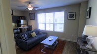 This single-floor space offers a comfortable and functional layout designed for everyday living. The living area serves as the heart of the home, centered around a tufted navy blue sofa accented with decorative throw pillows and paired with a modern two-tier coffee table. A warm, rich-toned area rug anchors the seating arrangement, adding texture and contrast to the hardwood-style flooring that runs throughout the space. Large windows dressed with white horizontal blinds allow abundant natural light to pour in, creating a bright and welcoming atmosphere. A ceiling fan with an integrated light fixture provides both air circulation and overhead lighting, contributing to the room's practical comfort. Wall art adds personality to the neutral-toned walls, which provide a clean and versatile backdrop throughout. A small dining or accent table is visible near the kitchen area, hinting at an open-concept flow between the living and dining spaces. The overall ambiance is contemporary and casual, with a neutral color palette punctuated by bold accent tones. The space is thoughtfully arranged to maximize both comfort and functionality within its single-level footprint.