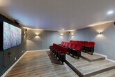 Located at 1501 Harvey Road in College Station, TX, this dedicated screening room delivers an authentic cinema experience within a well-designed, single-floor space. The room is arranged with rows of classic red-upholstered, theater-style seats mounted on black frames, organized across two raised tiers to ensure clear sightlines from every position. The tiered platform design elevates the rear rows above the front, mimicking the layout of a traditional movie theater.

The accent-painted front wall, finished in a deep charcoal tone, serves as the focal point of the room, anchoring a large wall-mounted flat-screen display ideal for presentations, screenings, or events. The remaining walls are finished in a neutral gray, contributing to a clean and focused viewing environment. Light wood-look flooring runs throughout, providing a warm contrast to the darker seating and feature wall.

Ambient lighting is provided by stylish dual-directional wall sconces positioned at intervals along the side and rear walls, casting soft upward and downward light to enhance the cinematic atmosphere without disrupting the screen. Recessed ceiling lights offer additional illumination when needed. The space is equipped with proper safety features, including a clearly marked exit door and fire extinguisher. The overall ambiance is polished, functional, and well-suited for immersive viewing experiences.