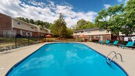 Located at 2525 Booker Creek Rd in Chapel Hill, North Carolina, this outdoor pool area is a well-appointed recreational amenity designed for relaxation and leisure. The centerpiece of the space is a generously sized swimming pool with a classic oval-rounded shape, filled with clear blue water and accessible via stainless steel entry rails. The pool's smooth concrete surround provides ample deck space for movement and socializing.

Lining the pool deck is a row of teal-colored lounge chairs, offering residents comfortable spots to sunbathe and unwind. A shaded seating area with a vibrant orange umbrella and patio furniture provides a welcoming retreat from direct sun. The entire pool area is enclosed by a sturdy black iron fence, ensuring both safety and security.

Adjacent to the pool is a single-story brick utility building with a pitched roof and a white entry door, likely housing pool equipment and support facilities. The surrounding landscape features mature deciduous trees that provide natural shade and a lush green backdrop, enhancing the open-air atmosphere. Under the expansive North Carolina sky, this pool area offers a refreshing and functional outdoor retreat within a well-maintained residential community.