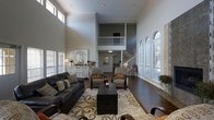 This impressive residence showcases a dramatic double-height living room that immediately commands attention upon entry. Soaring ceilings stretch well above the main floor, creating a grand sense of openness and volume. A classic white staircase with a traditional balustrade leads to an interior mezzanine overlook, adding architectural depth and visual interest to the space.

The living area is anchored by rich, dark hardwood flooring that extends throughout, providing a warm contrast to the soft grey walls and bright white trim. A series of elegant arched windows along one side of the room floods the space with abundant natural light, while additional clerestory windows near the roofline further enhance the bright, welcoming ambiance.

The layout is thoughtfully arranged, with a comfortable seating area centered on a patterned area rug. Classic crown molding and recessed lighting add refined finishing touches to the interior. A white vintage-style credenza and decorative wall art contribute to the home's timeless, transitional aesthetic.

Beyond the main living space, a glimpse into adjacent rooms suggests a well-connected floor plan with continued attention to detail. Overall, the home balances architectural grandeur with a warm, livable character, making it both visually striking and inviting.