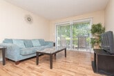 This single-floor, two-bedroom space offers a practical and cohesive layout designed for comfortable everyday living. The living area features warm hardwood-style flooring, neutral beige walls, and an open sightline into the dining area, where a classic dark wood table and chairs create a dedicated space for meals. A bar-height counter adjacent to the kitchen provides additional seating options.

The kitchen is equipped with dark upper cabinetry offering ample storage, speckled laminate countertops, a double-basin stainless steel sink, and black gas appliances. The light wood-tone flooring in the kitchen complements the cabinetry and creates a clean, functional workspace.

Both bedrooms are carpeted and finished with the same warm neutral palette. The primary bedroom accommodates a full-sized bed with a tufted upholstered headboard flanked by matching dark nightstands. The second bedroom features a similarly sized bed with bold patterned bedding and decorative wall art, adding personality to the space.

The bathroom includes a dark vanity with speckled countertop, an oval undermount sink, a well-lit mirror, and textured walls, maintaining the consistent design language found throughout the space. Overall, the layout is compact yet efficiently organized.