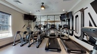 This fitness center is thoughtfully designed to accommodate a full range of workout needs within a well-organized, single-level space. The layout is divided into distinct functional zones, seamlessly connected through glass-paneled sliding doors that allow natural light to flow between areas and maintain visual openness throughout.

The cardio zone is anchored by a row of Landice treadmills and elliptical trainers, all positioned in front of floor-to-ceiling mirrors that visually expand the space. Multiple wall-mounted flat-screen televisions are positioned at eye level for convenient viewing during workouts. A ceiling fan provides additional air circulation, complementing the recessed lighting overhead.

The strength training area features a multi-function cable machine, adjustable bench press, free weights with a dedicated dumbbell rack, a plate-loaded weight tree, and a rowing machine. Large windows along one wall allow natural light to brighten this section while offering outdoor views.

Bold, large-scale typographic murals in black and white serve as a defining design element, giving the space a contemporary, energetic aesthetic. The light-toned flooring throughout provides a clean, cohesive look that ties both zones together, while the overall design balances functionality with modern visual appeal.