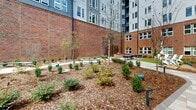Located at 5810 Stax St NW in Huntsville, Alabama, this thoughtfully designed outdoor courtyard is nestled within a contemporary multi-story building complex that combines brick, lap siding, and large-pane window facades. The courtyard is defined by a rich mix of architectural materials and lush landscaping, with winding paver pathways guiding visitors through mulched garden beds filled with ornamental shrubs, young trees, and native plantings. A standout feature is the sleek pergola seating area outfitted with comfortable lounge furniture, providing a shaded retreat for relaxation. Adjacent to it, a fully equipped outdoor kitchen with stainless steel grills and ample counter space makes alfresco dining and entertaining convenient. An open lawn area with vibrant artificial turf hosts an outdoor fitness station, complemented by classic Adirondack chairs positioned nearby for casual seating. Natural boulders are artfully integrated into the landscape, adding an organic contrast to the structured hardscaping. Glass-and-steel entry doors connect the courtyard seamlessly to the building's interior, ensuring easy access. The overall ambiance strikes a balance between urban sophistication and approachable outdoor living, offering a versatile community space ideal for recreation, socializing, and relaxation.