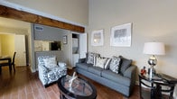 This two-floor space combines rustic charm with modern comfort, offering an inviting and well-appointed interior throughout. The living area immediately draws the eye upward with its soaring ceilings, creating an impressive sense of openness and volume. A distinctive exposed wood beam adds a warm architectural accent overhead, grounding the space with character and a touch of natural texture. The living room is thoughtfully furnished with a grey sofa, a coordinating patterned armchair, and a sleek glass-top coffee table, all resting on rich wood-toned flooring that flows throughout the main level. Framed wall art contributes to the polished, curated aesthetic. An open passage connects the living area to the kitchen, which features a raised breakfast bar with overhead cabinetry and modern appliances visible beyond. Adjacent to the kitchen, an in-unit laundry area adds practical convenience. The neutral grey and white color palette throughout creates a calm, cohesive ambiance, while the overall layout is designed to maximize both function and livability across two floors. The space strikes a balance between everyday practicality and an elevated, welcoming atmosphere.