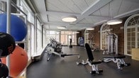 This fitness center combines classic architectural character with modern amenities to create a welcoming and functional workout environment. The space is defined by a striking contrast between exposed brick walls and crisp white coffered ceilings, lending a distinctive aesthetic that balances historic charm with contemporary design. Large, floor-to-ceiling windows line one side of the room, flooding the interior with abundant natural light and offering views of the surrounding exterior — an uplifting backdrop for any workout. Oversized circular pendant lights hang from the coffered ceiling, providing warm, even illumination throughout the space. The floor is finished with durable dark rubber matting, ideal for high-traffic fitness use. Equipment includes rowing machines, elliptical trainers, stationary bikes, a weight bench, and additional strength training apparatus, thoughtfully arranged to maximize floor space and ease of movement. Elegant arched windows with decorative mullions on the brick wall add a refined architectural touch, further enhancing the space's character. A water vending station is also available for convenience. This single-floor facility offers a spacious, light-filled setting well-suited for a comprehensive fitness routine.