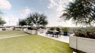 This distinctive single-floor space blends sophisticated interior design with impressive outdoor living. Inside, the dining area serves as a focal point, anchored by a striking round green marble table with an ornate pedestal base, surrounded by rattan-style chairs atop a woven area rug. Polished concrete floors run throughout, lending an industrial-modern character to the interior. White walls provide a clean backdrop for curated wall art, while a sliding barn door adds architectural charm and serves as a thoughtful design detail connecting interior spaces. Recessed lighting and track lighting ensure the interior is well-illuminated throughout.

The crowning feature of this space is the expansive rooftop terrace, which offers sweeping views of a city skyline. The terrace is thoughtfully designed with a combination of artificial turf and a paved entertaining area, creating distinct zones for relaxation and dining. Large white planter boxes filled with mature trees and lush shrubs provide privacy and a garden-like atmosphere. A built-in outdoor kitchen with a gas grill makes the terrace ideal for entertaining, while outdoor dining furniture offers a comfortable setting to take in the panoramic urban views. The overall ambiance is modern, curated, and effortlessly connected to the outdoors.
