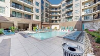 Located at 201 Jefferson St N in downtown Huntsville, Alabama, this amenity courtyard is a thoughtfully designed outdoor retreat nestled within a multi-story residential building. The centerpiece of the space is a generously sized resort-style swimming pool with crystal-clear blue water, flanked by rows of vibrant blue lounge chairs ideal for sunbathing. A shallow tanning ledge with in-water chaise lounges and water jets provides a distinctive leisure feature, while private poolside cabanas with teal curtains offer shaded retreats for relaxation. The surrounding deck is finished in large-format tile, providing a clean, modern aesthetic throughout. A covered pergola with string lights and ceiling fans anchors a secondary lounge and dining area, framed by tall columnar evergreen trees and lush tropical plantings that add a rich layer of greenery. Thoughtfully curated landscaping, including specimen trees, ornamental shrubs, river rock accents, and oversized planter urns, soften the brick and panel exterior of the building. Large floor-to-ceiling glass windows reveal an interior fitness center stocked with equipment and exercise balls. The entire courtyard benefits from abundant natural sunlight and is enclosed by the building's multi-story facade with private balconies overlooking the space.