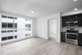 This single-floor unit showcases a contemporary design characterized by clean lines, a neutral white color palette, and light hardwood flooring throughout. The open-concept living area is anchored by large, floor-to-ceiling windows that flood the space with natural light while offering views of the surrounding building exterior. The layout flows seamlessly from the main living area toward a separate room, accessible through a clean white-framed doorway.

The kitchen is thoughtfully designed with high-gloss gray cabinetry extending to the ceiling, providing ample storage. Stainless steel appliances, including a full-size refrigerator and dishwasher, complement the light countertops and a functional sink. Recessed ceiling lighting enhances visibility and adds a polished finish to this area.

The bathroom maintains the unit's minimalist aesthetic, featuring light wood-tone flooring, white walls, and chrome hardware accents. A vanity with a stone-look countertop adds a refined touch to the otherwise understated space.

Throughout the unit, white walls and smooth ceilings create a bright, airy ambiance. An electrical panel is discretely integrated into the living area wall. Overall, the space presents a well-organized, modern environment suited to efficient, contemporary living.
