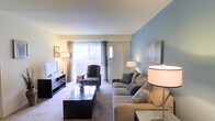 This single-floor residence offers a thoughtfully designed living space that balances comfort and modern style. The living room serves as the centerpiece of the home, showcasing a soothing two-tone color scheme with soft beige walls complemented by a striking blue accent wall that adds depth and character to the room. Plush neutral carpeting runs throughout, contributing to a warm and inviting atmosphere. A large sliding glass door flanked by elegant curtains floods the space with an abundance of natural light, creating an airy, open feel and providing a direct connection to the outdoors. The living area is generously proportioned, comfortably accommodating a full seating arrangement that includes a spacious sofa adorned with decorative pillows, a patterned armchair, and a sleek black lacquer coffee table. A contemporary media console along the wall provides ample storage and display space. The layout flows efficiently, making practical use of every area. Recessed and floor lamp lighting options allow for flexible ambiance throughout the day and evening. Overall, the space presents a clean, well-maintained interior that is move-in ready and suitable for a variety of lifestyle needs.